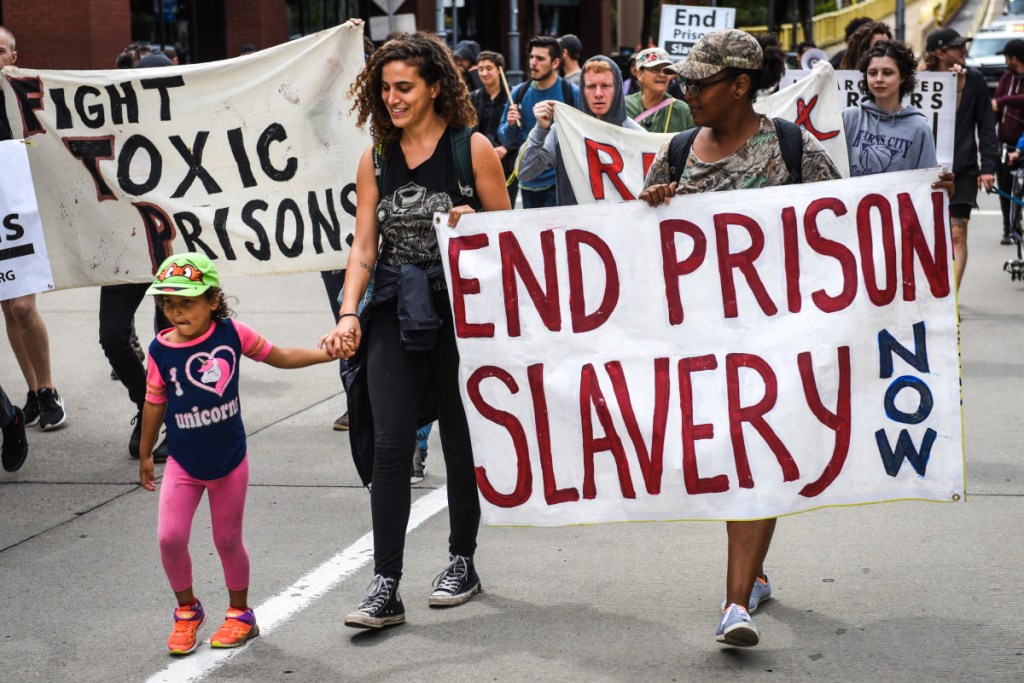 Two adults and a child march during a protest, with banners reading "Fight Toxic Prisons" and "End Prison Slavery Now."