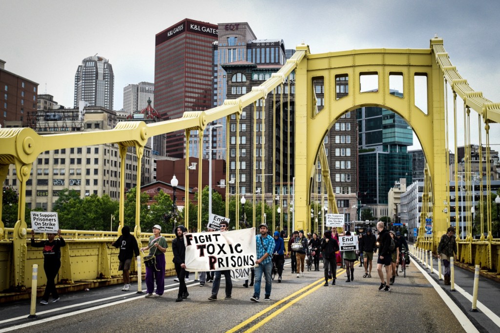 A protest march crosses a bridge, carrying a banner that reads, "Fight Toxic Prisons."